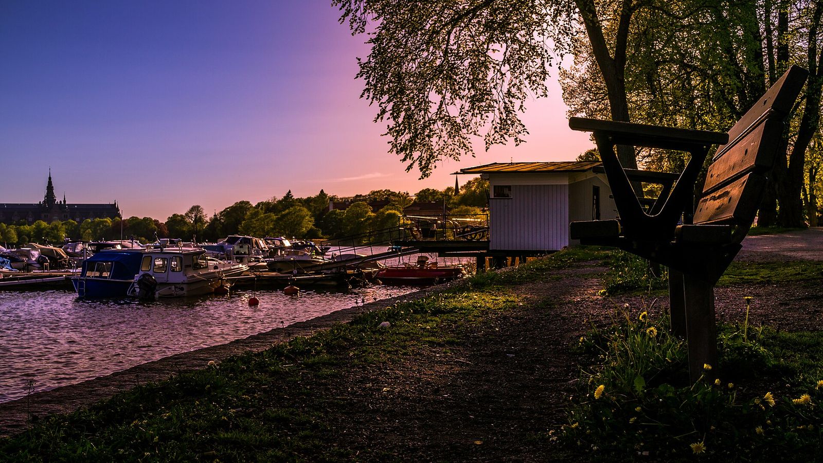 Småbåtshamn i skymningen med förtöjda båtar och en bänk under träden vid stranden