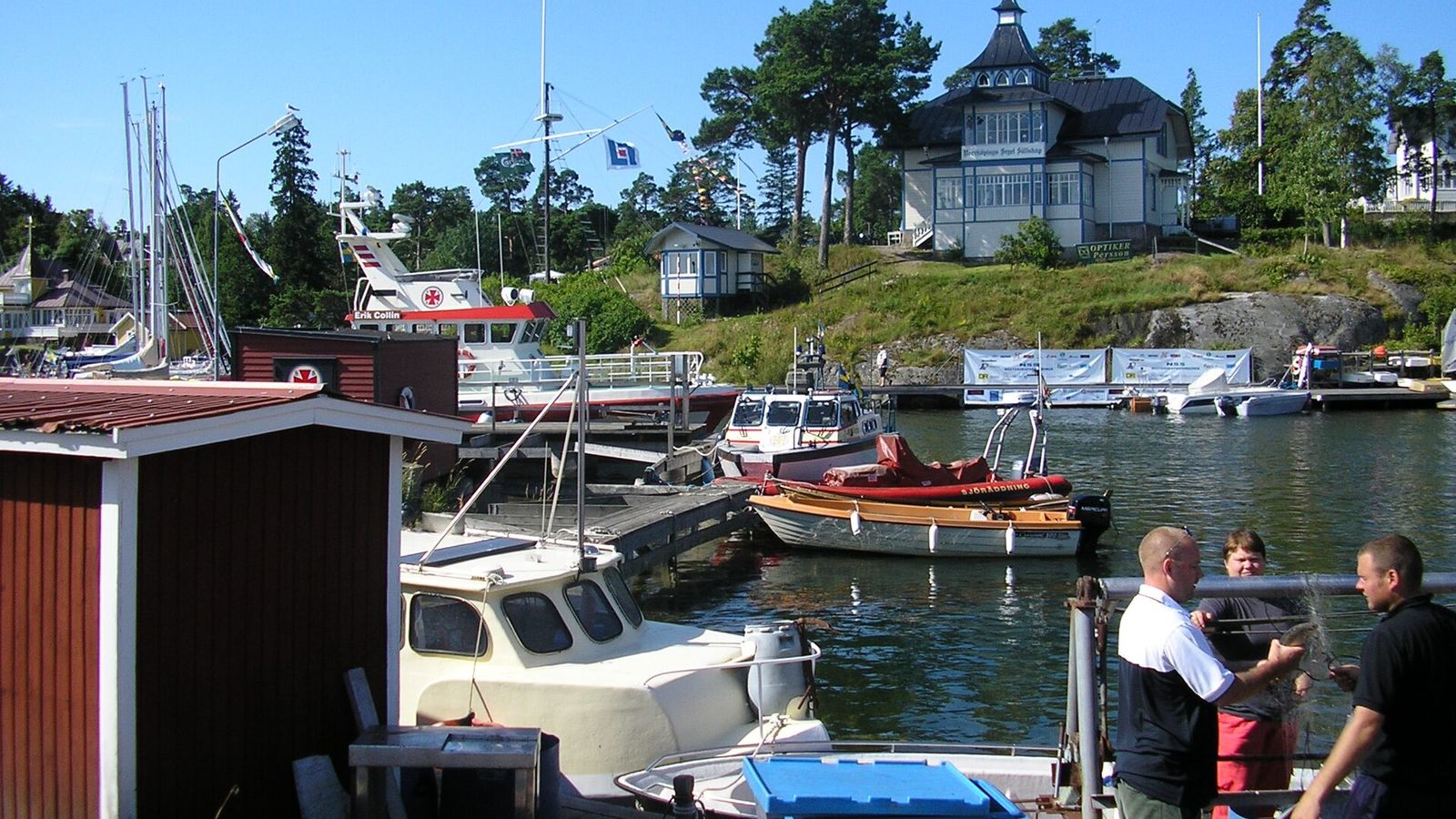 Sommardag i Arkösunds hamn med folk på bryggan, småbåtar och sjöbodar